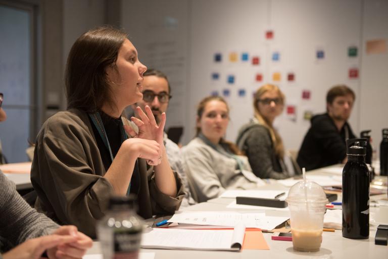 A person in the left foreground sits at a long table, talking and gesturing with their hands. Sheets of paper, pens and drinks sit on the table. Four other figures listen intently. Partially obscured.