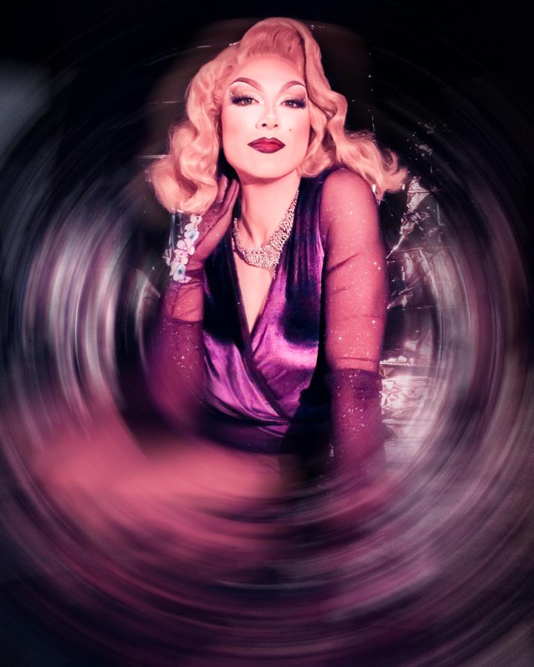 Drag queen with shoulder-length blonde hair wearing silver jewelry and a purple dress poses in front of a silver background. Partially obscured.