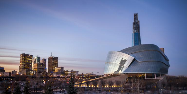 The exterior of the Canadian Museum for Human Rights, seen in the sunset against the city skyline. Partially obscured.