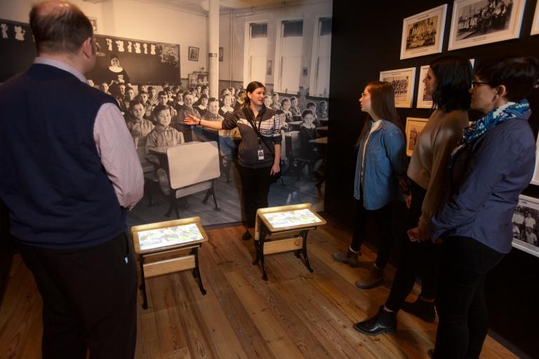 A visitor looks at a gallery niche on residential schools at the Canadian Museum for Human Rights. Two small desks sit in the middle of the niche. A large image of Indigenous students sitting at desks at a residential school is on the back wall of the niche, with images and artifacts on the side walls. Partially obscured.