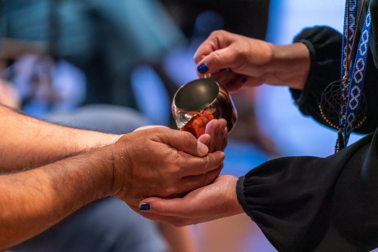 A person pours water from a copper cup into the cupped hands of another person. Partially obscured.