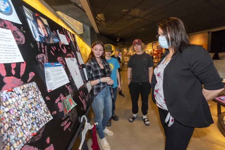 Three students stand in front of a poster board, placed on a table. A museum visitor stands in front of them looking at the poster board. Partially obscured.