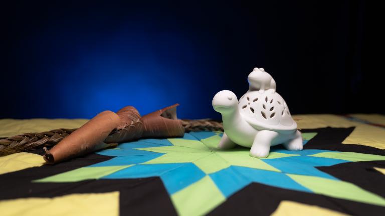 A white figure of a turtle with a frog on its back sitting on top of a colourful star blanket. Partially obscured.