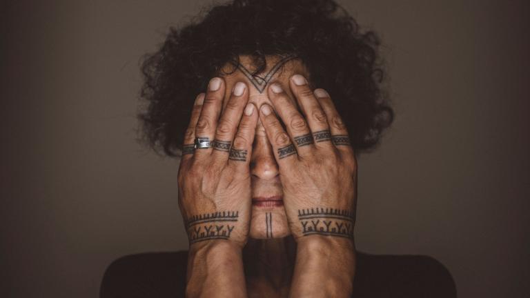 Aaju Peter covers her eyes and cheeks with her bare hands. Tattoos are visible on her forehead, chin, fingers and wrists. Her hands and the visible parts of her face are illuminated and are framed by her dark curly hair and angular shoulders. Partially obscured.