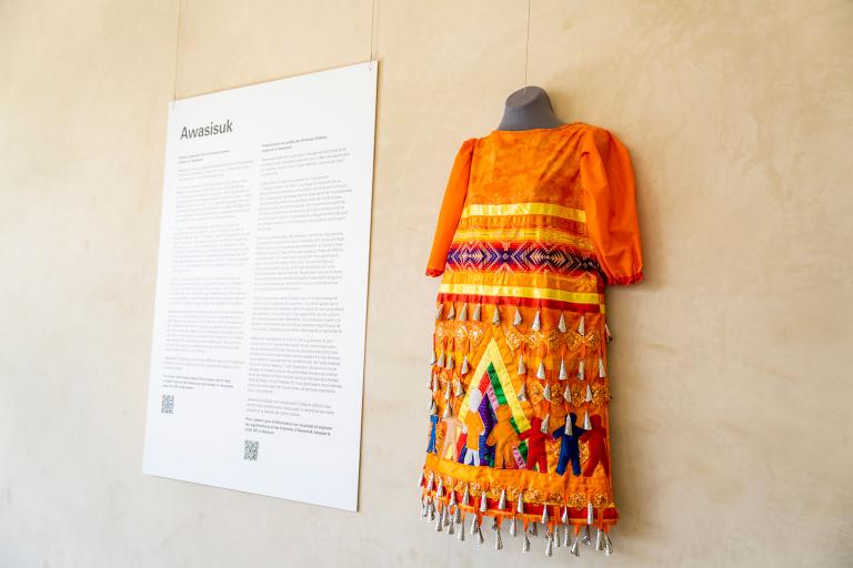 An orange jingle dress hangs beside a text panel. Jingles, or small silver cone-shaped pendants, are visible, as well as coloured ribbons and human figures in various colours and sizes. These human figures are holding hands inside a multicoloured tipi.