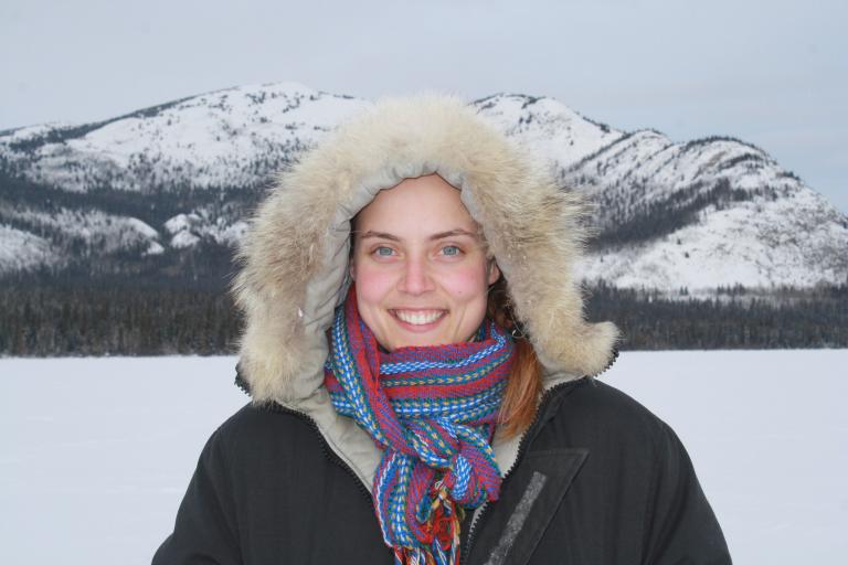 Smiling female wearing a fur-lined parka posing in front of a mountainous terrain. Partially obscured.