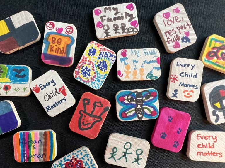 Small decorated wood tiles sit on a black backdrop. Some display images of families, others show messages like "Every child matters," "Be kind" and "Human is Human." Partially obscured.