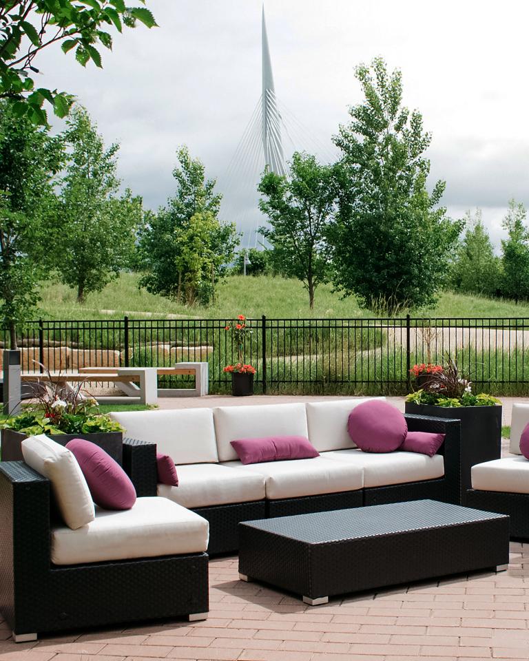 Couches and chairs sit in a fenced patio. Small trees and tall green prairie grass surrounds the patio. In the background, the spire from Esplanade Riel rises above the trees. Partially obscured.