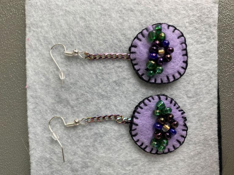 A pair of light purple felt earrings with dark purple sewn borders. In the middle of the earrings are beaded flowers. Partially obscured.