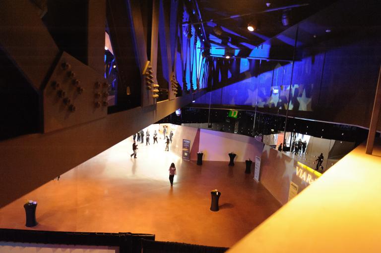 Several people stroll through a great open hall. They are viewed from a ramp above. The ramp’s metal structure and rivets are visible on the left, while on the right a gleaming mirrored wall reflects the people below plus vivid indigo lighting and white stars. Partially obscured.