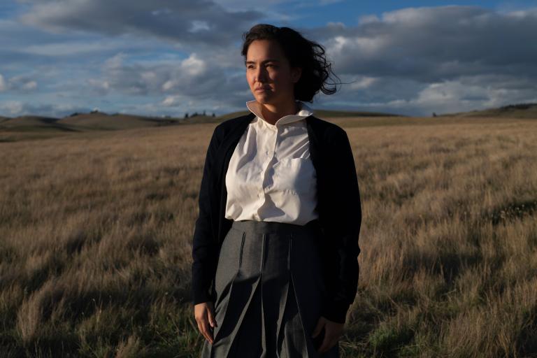 An Indigenous woman stands in a field of prairie grasses. There is a blue sky with grey clouds behind her as the sun shines on her. Partially obscured.