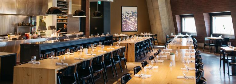 The dining area in a restaurant with long tables and a peek into the kitchen. Partially obscured.