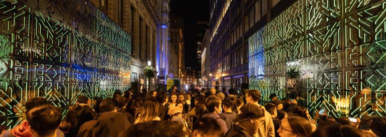 Many people stand between two intricately patterned joined mirrors that reflect green lights and a throng of onlookers. Partially obscured.
