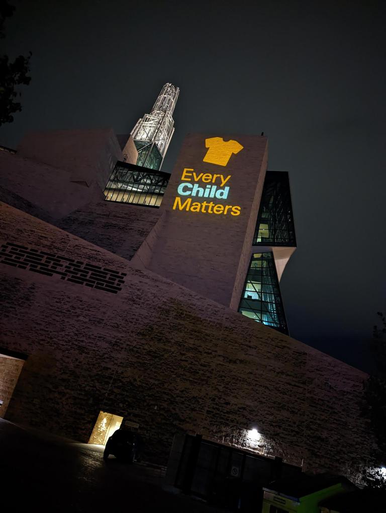 An image of an orange shirt with the text Every Child Matters is projected on an exterior wall of the Museum. Partially obscured.