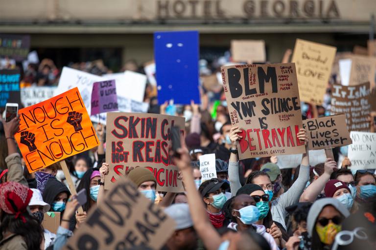 A large group of people holding signs with slogans such as “Black Lives Matter,” “Enough is Enough” and “Your Silence is Betrayal.” Partially obscured.