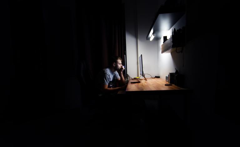 A white man with short hair sits alone at a brightly lit desk in a dark room, staring at a computer monitor. Partially obscured.