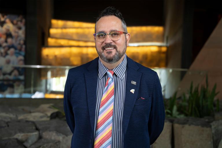Walter Cassidy stands in front of the glowing alabaster ramps at the Canadian Museum for Human Rights. On his suit lapel, a small pin reads "Queer." Partially obscured.