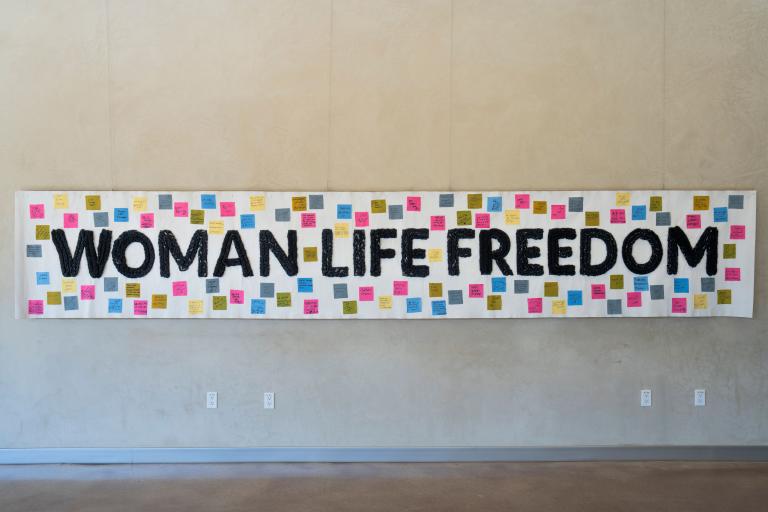 A white banner hangs on a wall, bearing the words "Woman, Life, Freedom" in black. Small coloured squares with text are attached to the banner around the text. Partially obscured.