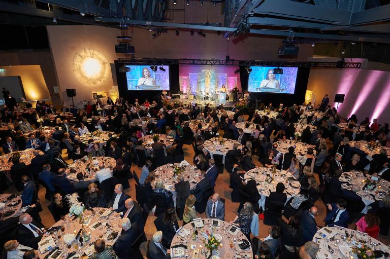 In a large hall, hundreds of diners sit at round tables while a woman speaks at a podium. Partially obscured.