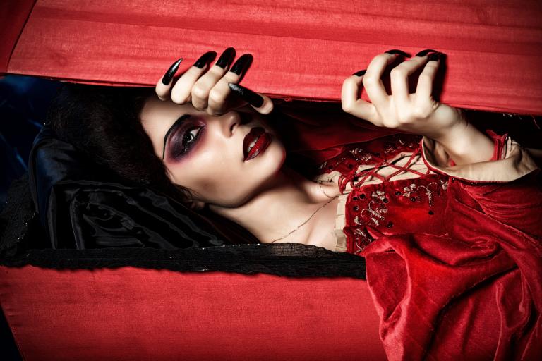 A vampire dressed in a velvety red dress peeks through a red wooden box with her long, black nails. Partially obscured.