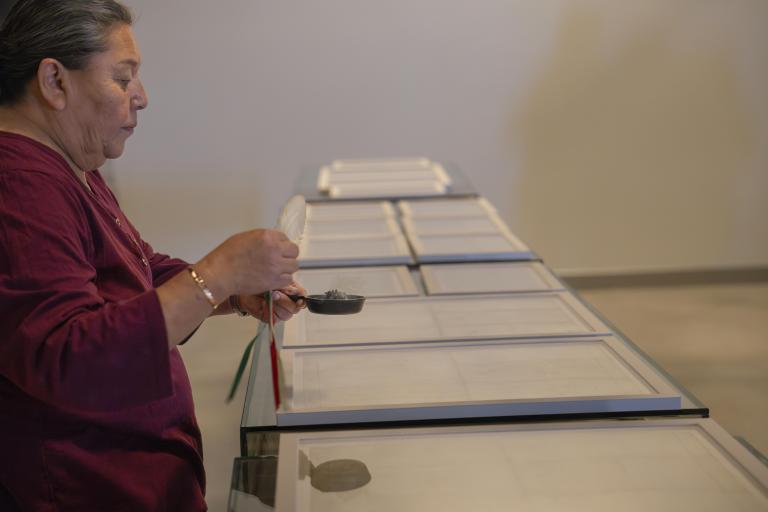 Framed documents sit on a table as Elder Hazel Copenace smudges them, holding a smudge bowl in her left hand and an eagle feather in her right.