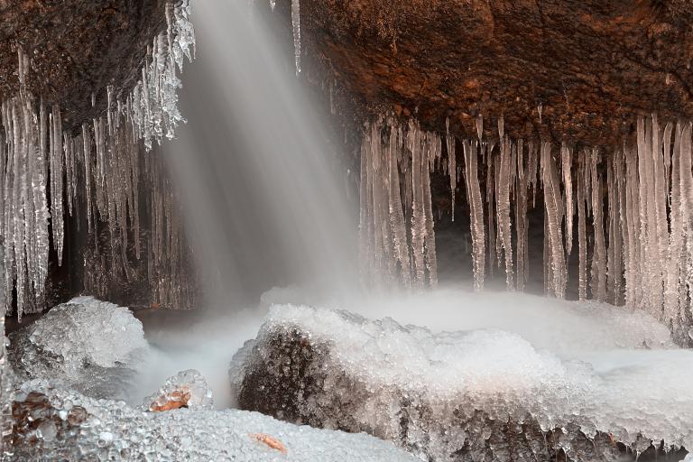 Water sprays onto rock formations from an unseen opening above, causing a dramatic array of icicles and mounds of ice on boulders. Partially obscured.