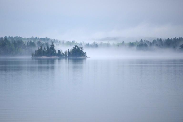 Mist rises off a large, calm lake surrounded by an evergreen forest. Partially obscured.
