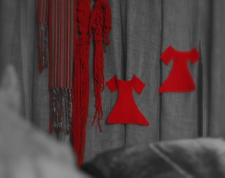 Two bright red felt dresses are pinned to a grey background next to braided red yarn and a traditional Métis sash. Partially obscured.