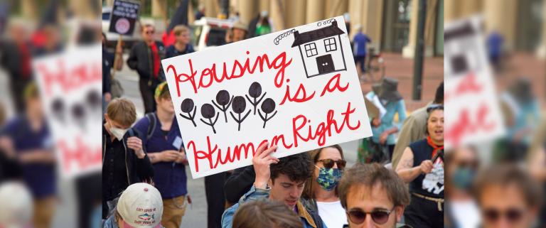 A group of people walking down a street. One person holds a sign that reads: “Housing is a human right.” Partially obscured.