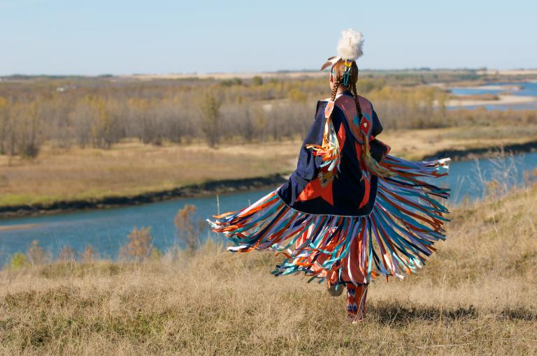 A Indigenous dancer in brightly coloured Fancy Shawl regalia beside a river. Partially obscured.