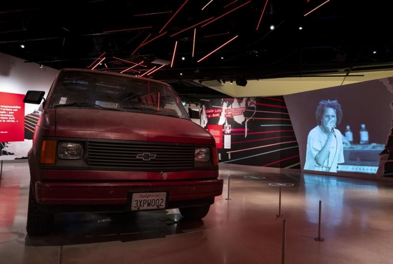 Red Chevrolet van in an exhibition space with video by Rage Against the Machine playing in the background. Partially obscured.