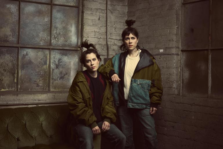 Two people with long hair in high ponytails wearing winter coats pose in the corner of a grey-painted brick room. Partially obscured.