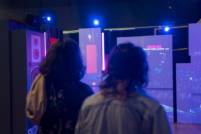 Two visitors, seen from behind, view a series of exhibition panels that show a large crowd at a rock concert. Partially obscured.