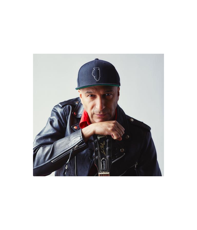 Tom Morello looks intently at the camera while leaning on his hand, which holds a guitar by the neck. He is wearing a black leather jacket, red collared shirt and baseball hat. Partially obscured.