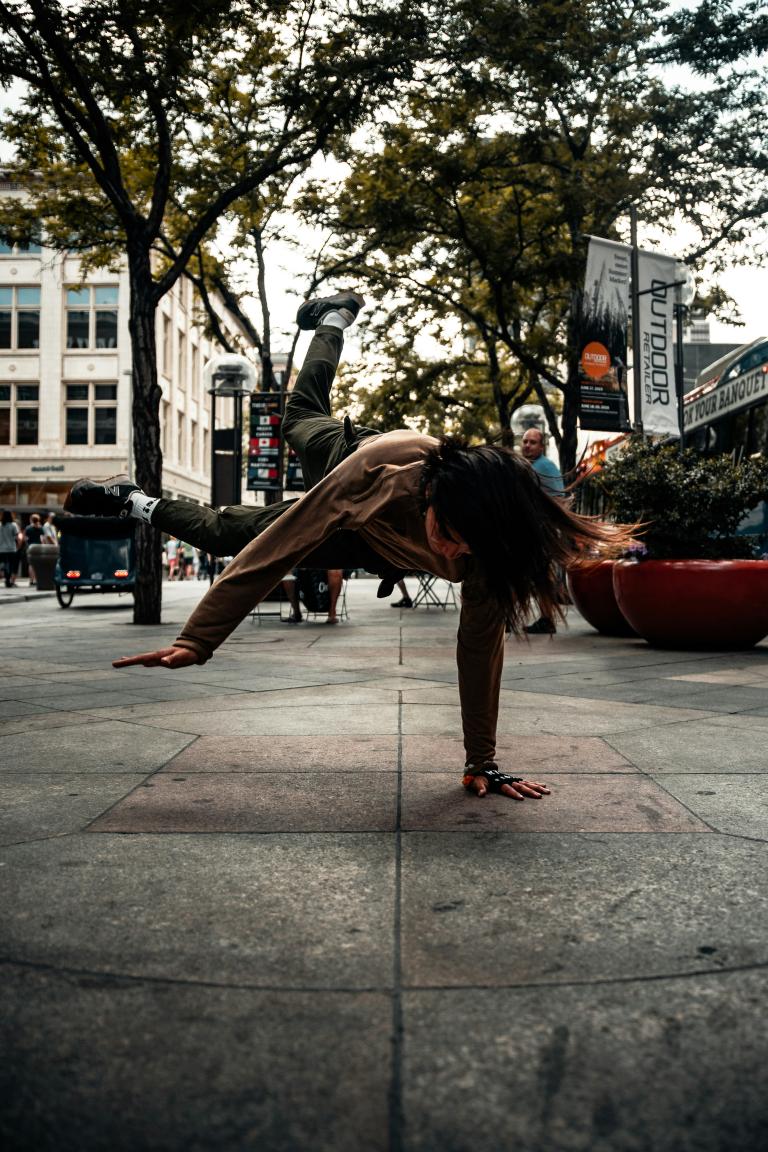 A woman in a grey jacket and darker grey pants is breakdancing on a sidewalk during daytime. Trees and buildings are in the background. Partially obscured.