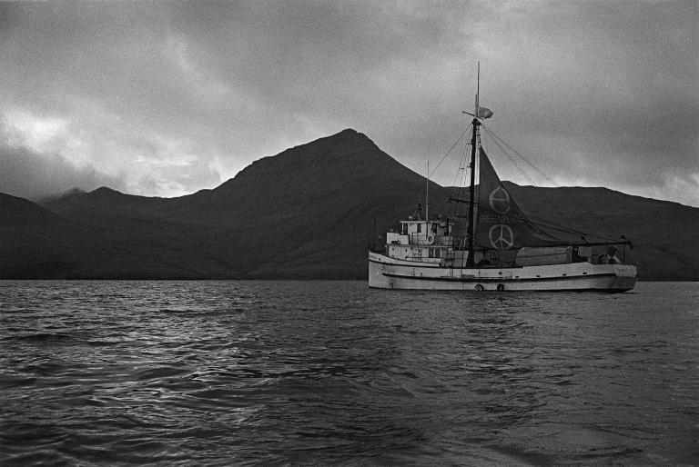 A boat in the water with mountains and clouds in the background. Its sail is decorated with large peace signs. Partially obscured.