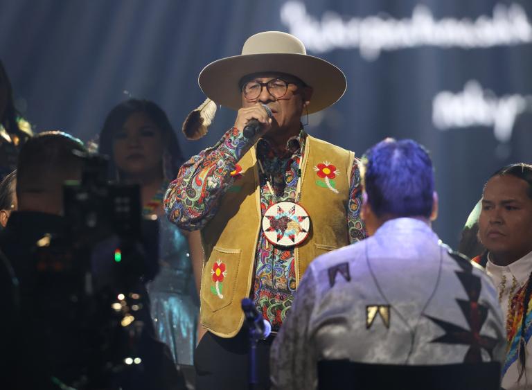 Close-up on a group of people wearing beaded jackets and vests performing on stage. In the centreer, a man wearing glasses, a hat, and a large, beaded medallion sings into a microphone. Partially obscured.