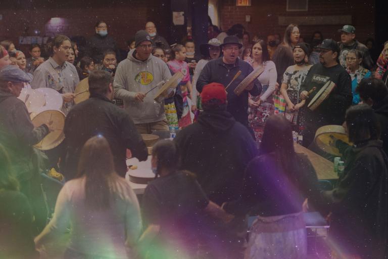 A group of men drum and sing in a circle. They are tightly surrounded by a large and diverse crowd of people all singing along. Partially obscured.