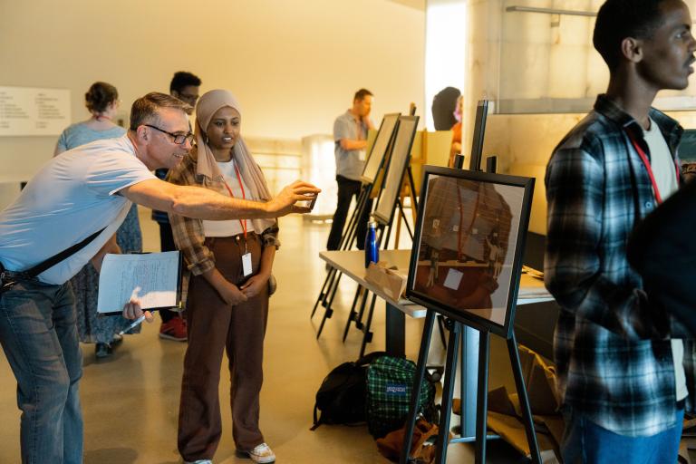 Students in a museum gallery display their work on easels. The focus is on one student talking with an adult who is smiling and taking a photograph with their phone of the student’s work. The artwork shows four people in a hallway with four doors, each with a sign on it. Partially obscured.