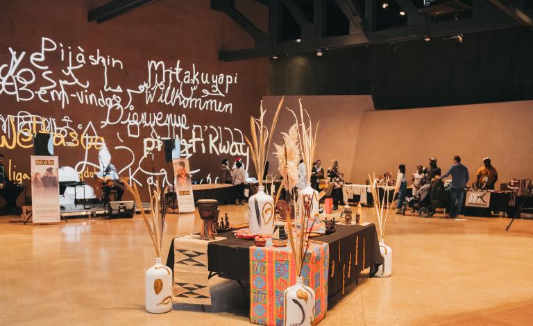 A scattering of people are in a room where kiosks are placed around it. In the centre of the room, there is a long table adorned with vibrant tablecloths and vases filled with long, dry plants. Partially obscured.