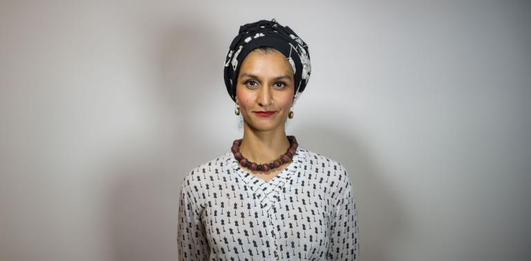 A Pakistani woman, Anam Abbas, in a black and white headwrap, red lipstick, chunky brown collar necklace and black-and-white blouse looks forward with a bemused expression. Partially obscured.