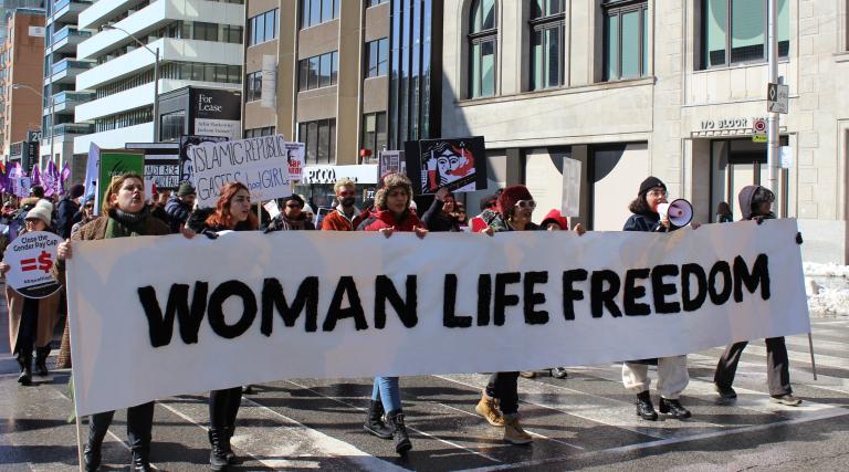 Six marchers carry a long white banner with black capital letters that read WOMAN LIFE FREEDOM down a Toronto city street. Behind them, a parade of women carry other protest signs for international Woman's Day. Partially obscured.