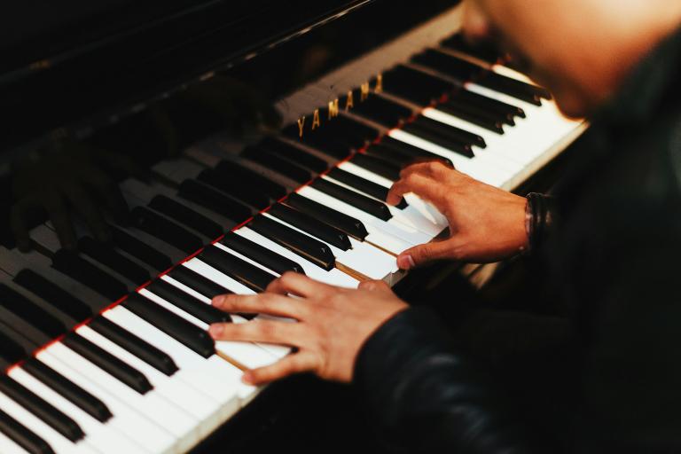Two hands play the keys of a gleaming black Yamaha piano. The pianist, barely shown, wears a long-sleeved black top. Partially obscured.