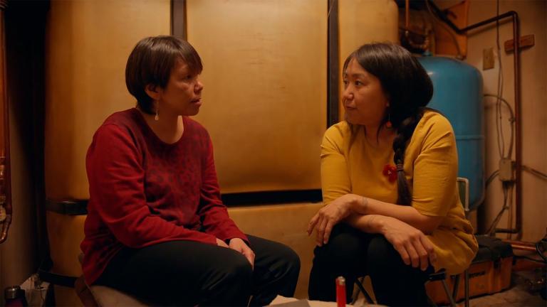 Two Inuit women sit in a dim room facing one another in intimate conversation. One is wearing a red sweater, the other a yellow sweater. Partially obscured.