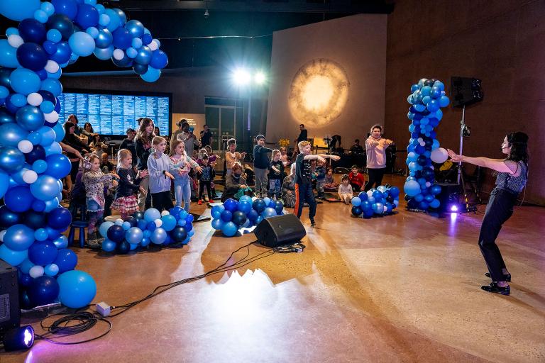 Kids dancing in a hall filled with blue balloons. Partially obscured.