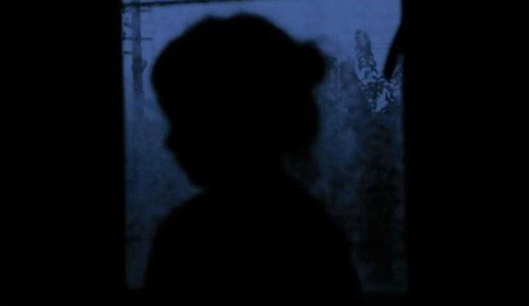A dark image of a young child’s silhouette against a window through which are seen blurred trees and a power pole. The light is a purplish tone of twilight. Partially obscured.