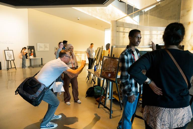 In a museum gallery, students exhibit projects on easels. Two students speak with adults about their work. Partially obscured.