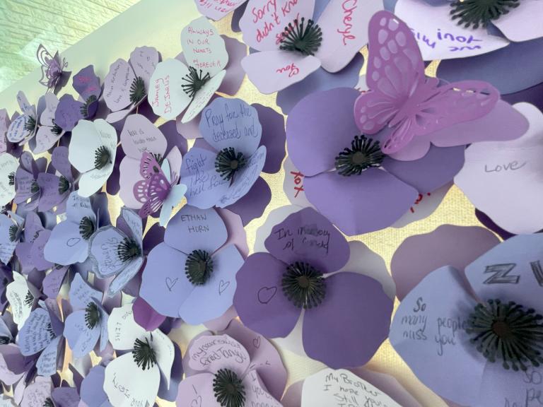 Purple poppies made of paper with names and messages from community members and family of those who have died as a result of a poisoned drug supply. Partially obscured.