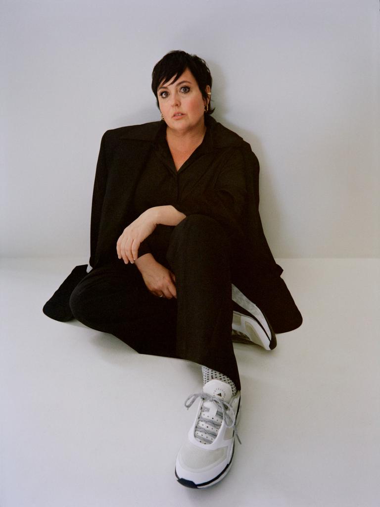 A woman with short black hair and wearing black clothes is sitting with her left arm bent across her knee and right arm by her side.