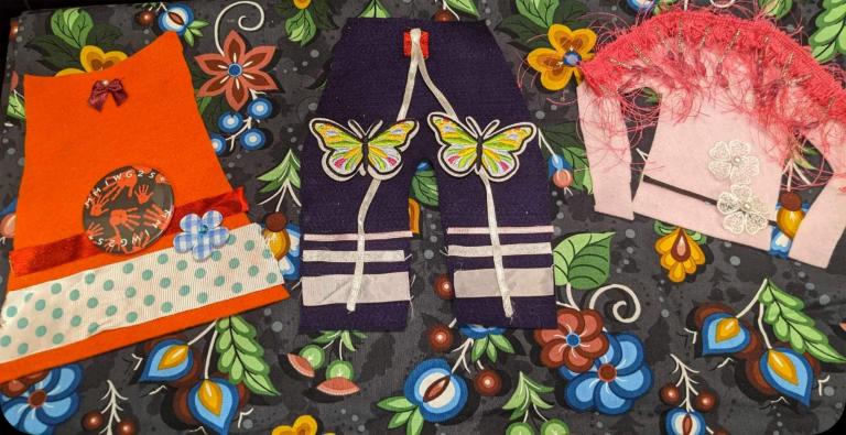 Pieces of crafted Indigenous- style clothing, including an orange shirt, pair of pants and made from colourful fabric and ribbon is are laid against a piece of craft paper adorned with flowers and berries. The clothing is includes an orange shirt, a pair of pants with ribbons and a pink tunic decorated with colourful fabric, appliqués and ribbons. The tunic has with a feathered shawl on the shoulders. Partially obscured.